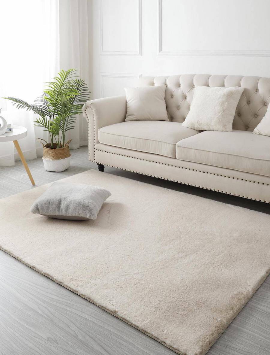 Living de estilo clásico con un sofá capitoné color crema con cojines en la misma tonalidad. Frente a él hay una alfombra de piel sintética color crema con un cojín gris encima.