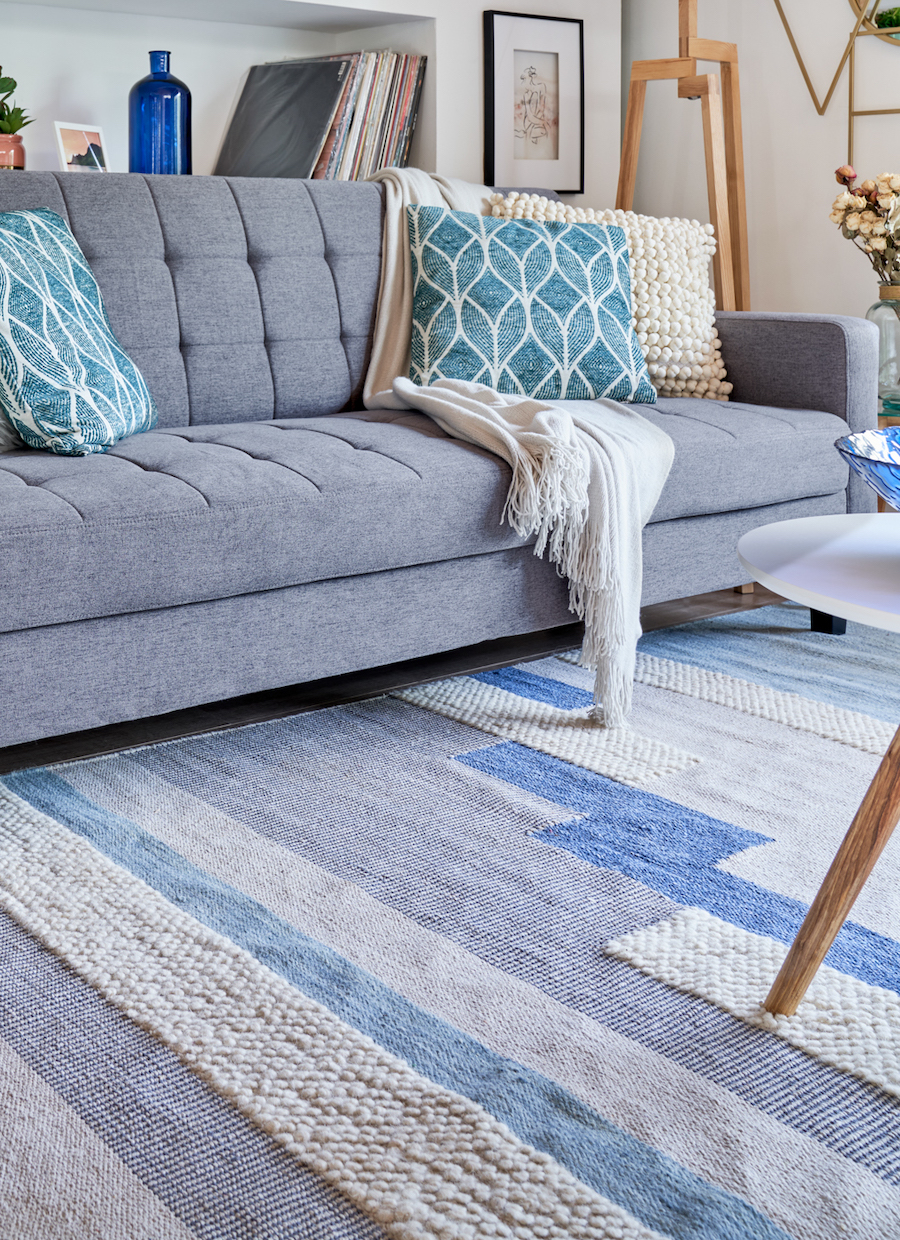 Detalle de un living con un sofá capitoné moderno gris con cojines crema y calipso junto a una alfombra con diseños geométricos en tonos azulados.