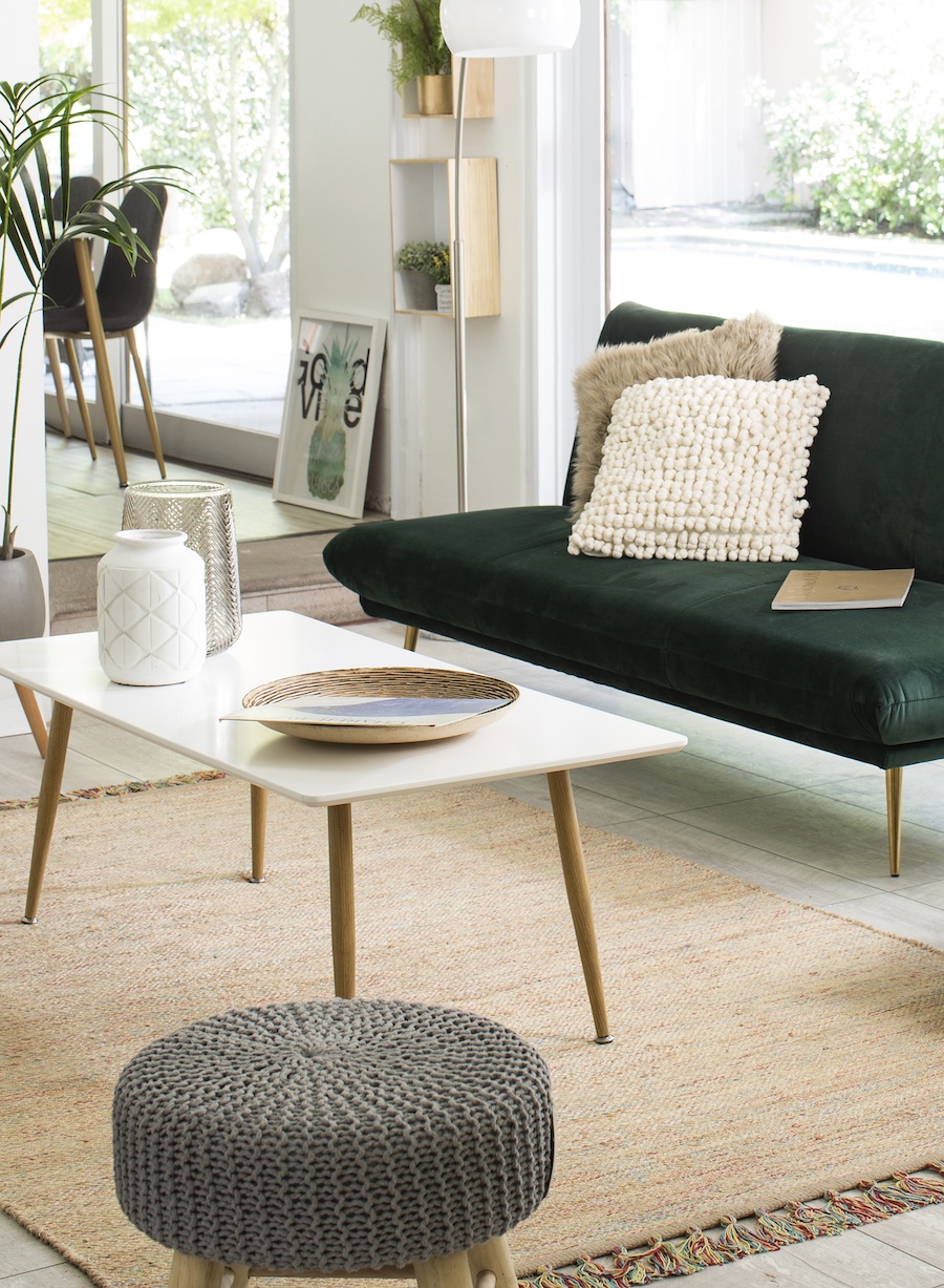 Sala de estar de muros blancos y un tipo de alfombra de yute. Sobre ella hay una mesa de centro de patas alargadas de madera y superficie blanca. Junto a ella, un futón verde oscuro con cojines en tonos crema.