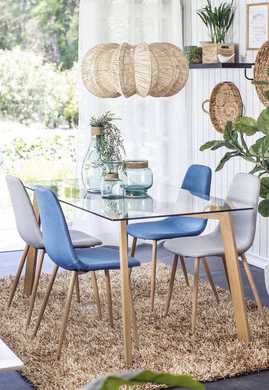 Comedor estilo playero con mesa de vidrio y sillas estilo eames gris y azul. Sobre la mesa cuelga una lámpara de techo de ratán, escultórica. Bajo la mesa hay una alfombra shaggy café claro.