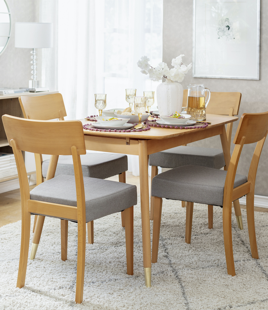 Comedor de madera clara con una mesa rectangular y cuatro sillas con tapiz gris. La mesa está puesta con vajilla blanca y copas de vidrio. Bajo la mesa hay una alfombra blanca con líneas grises