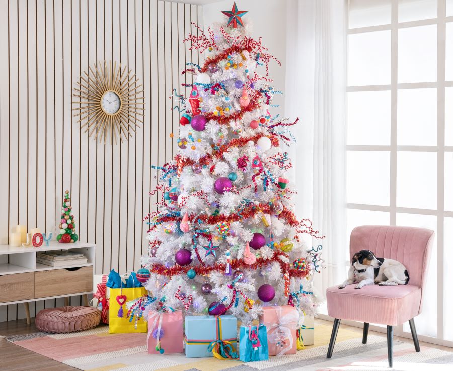 Living moderno con árbol de navidad blanco y adornos de muchos colores como celeste, rosado, amarillo. Lo rodea una guirnalda roja. Bajo el árbol hay regalos envueltos, y al lado un sitial rosado con un perro sobre él. Pared con revestimiento de madera