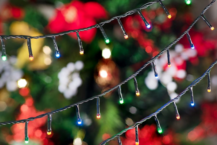 Detalle de tres tiras de guirnaldas de luces de navidad, de colores, encendidas. Atrás se ve un árbol de navidad con adornos rojos, blancos y dorados