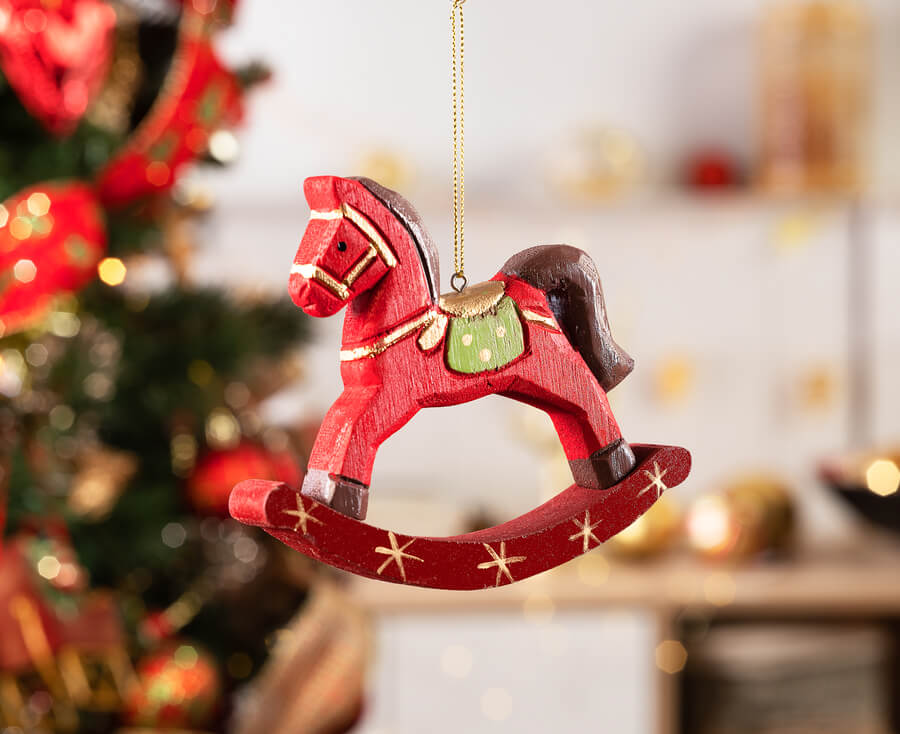 Detalle de un ornamento o decoración navideña de un caballo rojo, colgado con un hilo dorado. Atrás se ve un árbol de navidad.