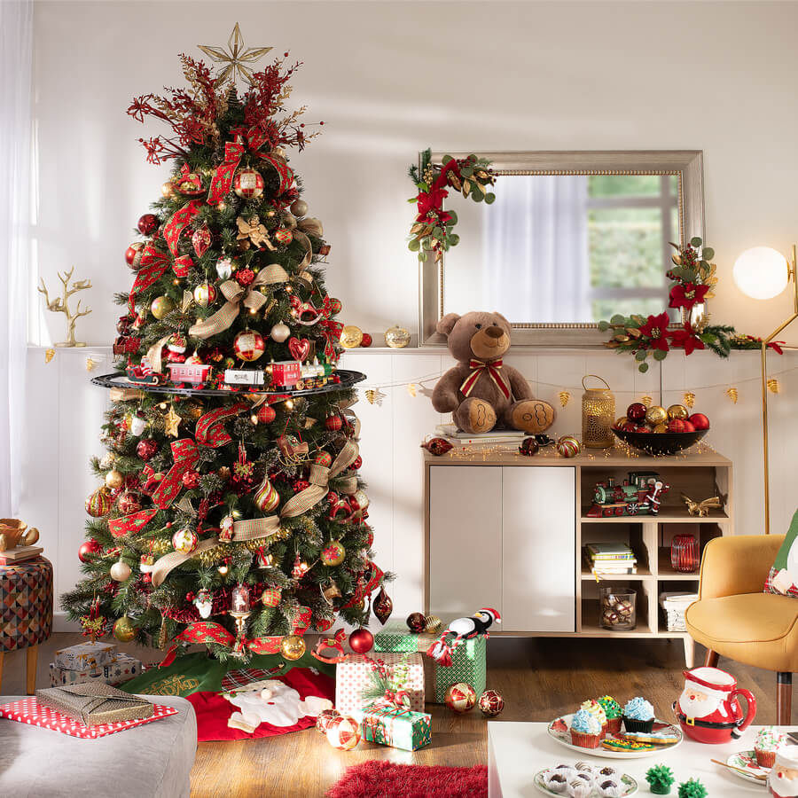 Gran árbol de navidad en un living. Tiene adornos rojos y dorados, con un tren. Regalos envueltos en el suelo. Junto al árbol hay un mueble de madera, con una puerta blanca y repisas. En el muro hay un revestimiento blanco y sobre él hay un cuadro
