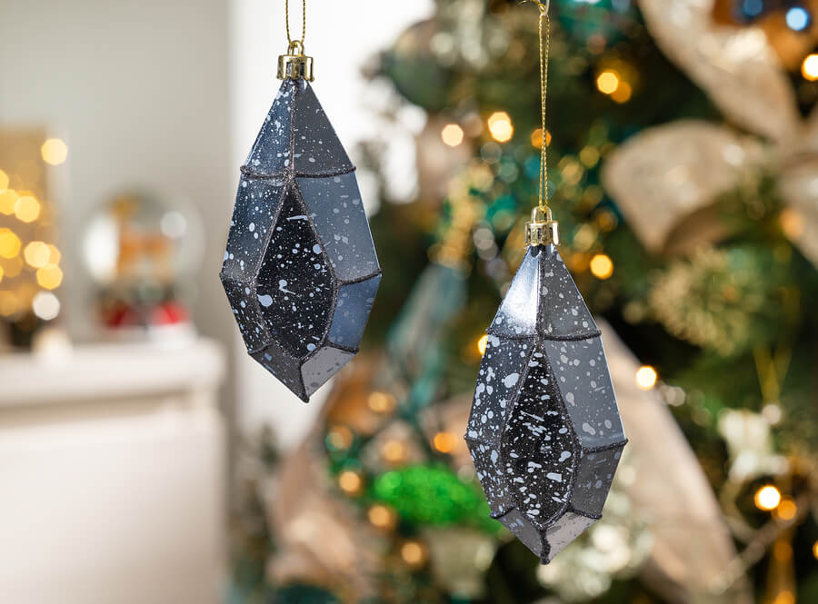 Detalle de dos ornamentos de navidad o adornos para árbol, de color azul y forma de lágrima geométrica. Tienen un hilo dorado y pintura jaspeada. Atrás se ve un árbol de navidad con adornos y las luces encendidas-