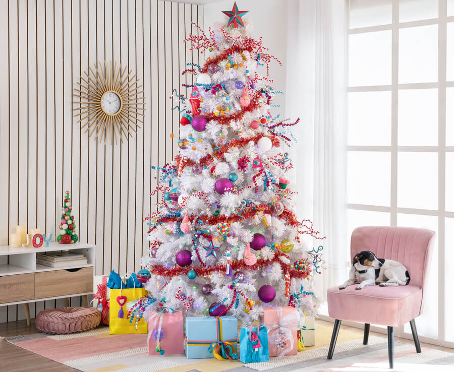 Árbol de navidad blanco, con decoraciones de colores, en un living. Tiene adornos rojos, rosados, celestes, amarillos y morados. Bajo él hay regalos envueltos con papeles de colores. Al lado hay un sitial rosado y en el suelo una alfombra.