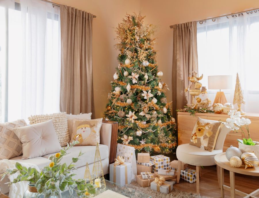 Living con un gran árbol de Navidad al centro, en tonos dorado. Sofá beige con cojines navideños, regalos envueltos en el suelo, un sitial y un mueble de madera bajo la ventana.