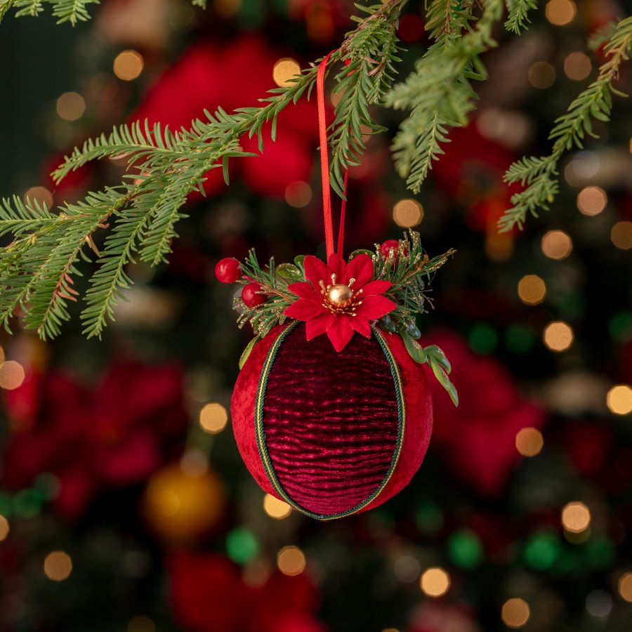 Detalle de un ornamento de Navidad redondo, forrado en tela aterciopelada en tonos rojos y burdeos, con una pequeña flor y hojas en la parte superior.