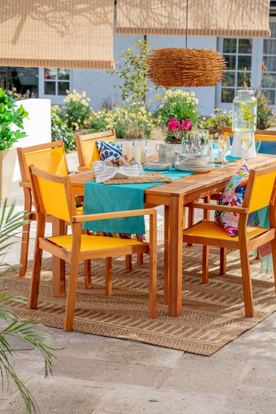 Terraza con mesa de comedor de madera, con 6 sillas ccon base y respaldo de tela color amarillo. Pesa puesta con vajilla blanca sobre un camino de mesa turquesa. Sobre la mesa hay una lámpara de techo redonda, de mimbre. Alfombra de sisal café