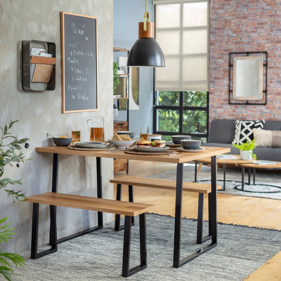 Comedor de estilo industrial junto a una pared gris. Bajo la mesa y banquetas hay una alfombra gris sobre un piso de madera. Atrás se ve un living de estilo industrial y un muro de ladrillos.