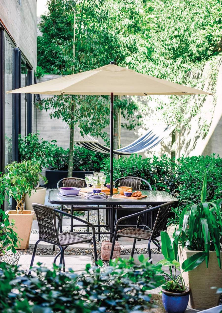 Jardín seco junto a una terraza con mesa y cuatro sillas. Tiene un quitasol beige y atrás se ve una hamaca azul con blanco.