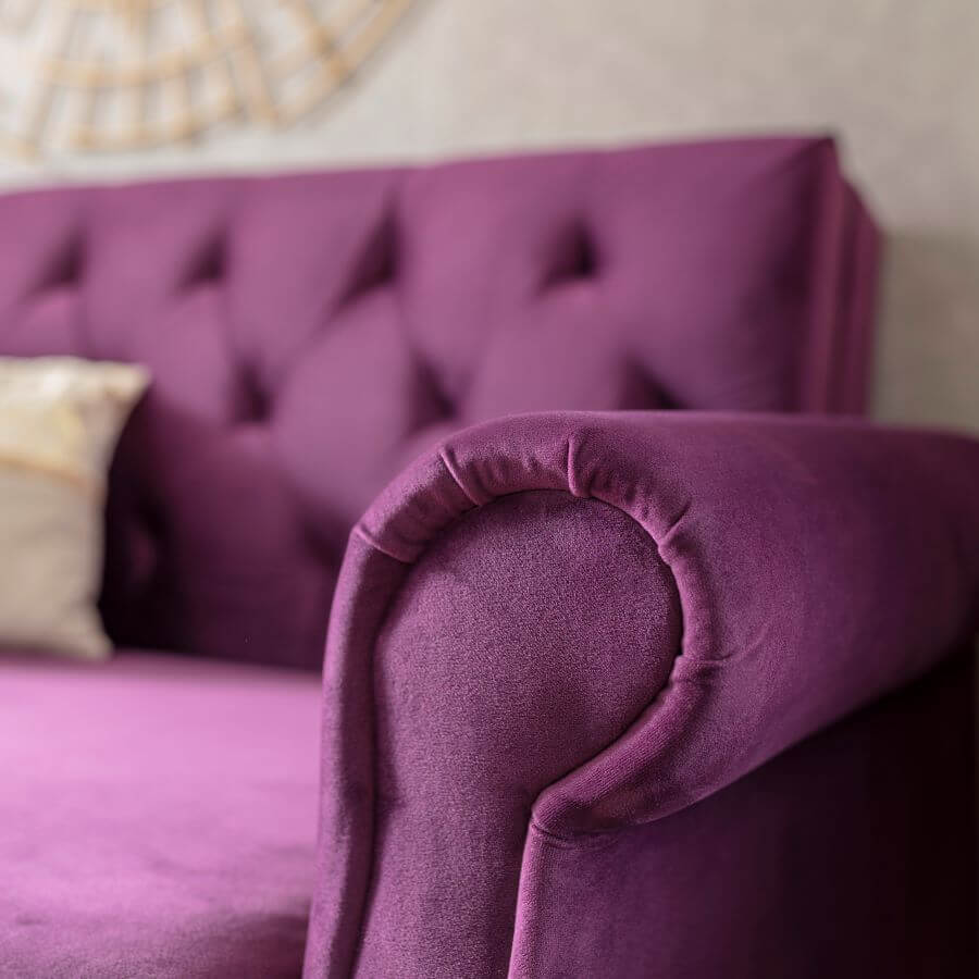 Detalle de sillón de terciopelo color morado, con respaldo capitoné. Se ve parte de un cojín de color beige y gris.