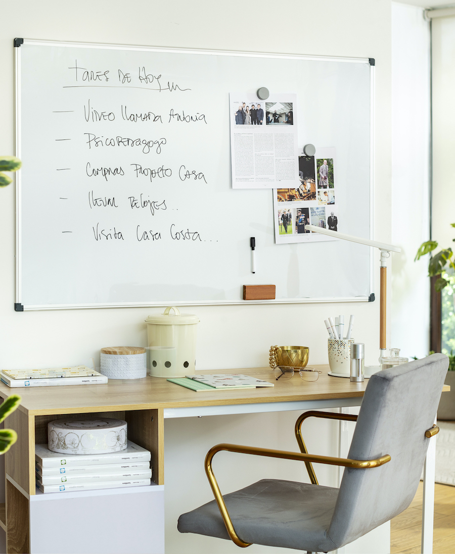 Espacio de trabajo con un escritorio de madera, una silla de terciopelo gris y detalles dorados. En la pared sobre el escritorio hay una pizarra blanca con tareas y papeles.