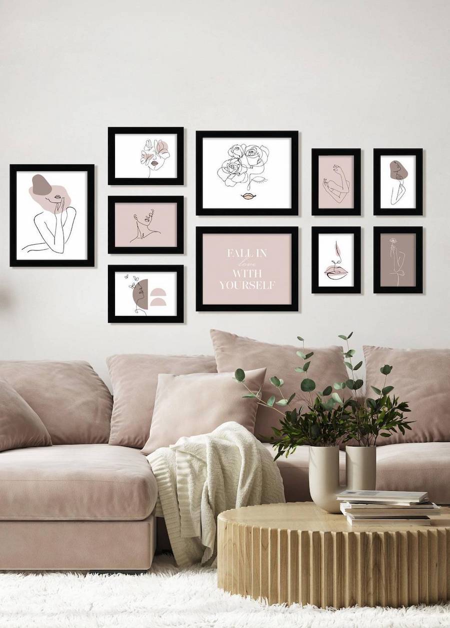 Sala de estar con un muro galería con ilustraciones en tonos rosado pastel con marcos negros, un sofá rosado con cojines del mismo color y una mesa de centro de madera clara y redonda.