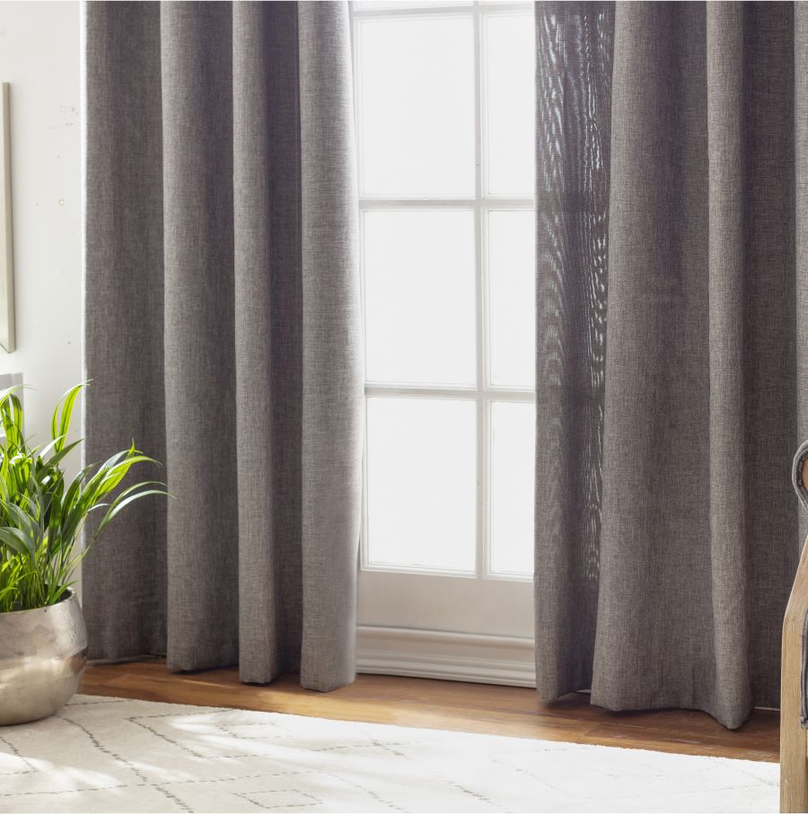 Acercamiento de un ventanal con marco de madera blanco, que está tapado casi por completo por una cortina de tela gris. A un costado se alcanza a ver una planta en un macetero plateado. Piso de madera con una alfombra blanca