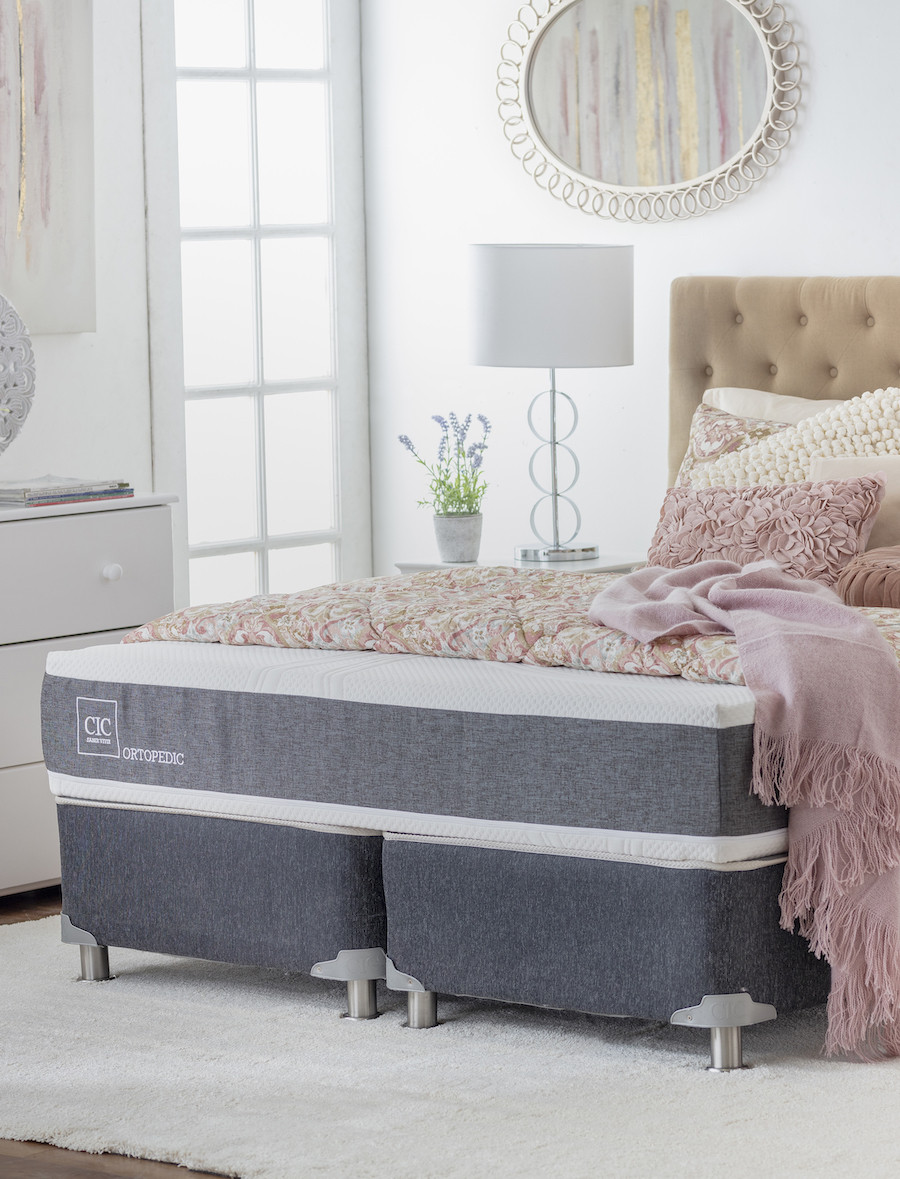 Cama de dos plazas con respaldo capitoné beige, colchón y ropa de cama de flores encima. También hay cojines en tonos rosado pastel.