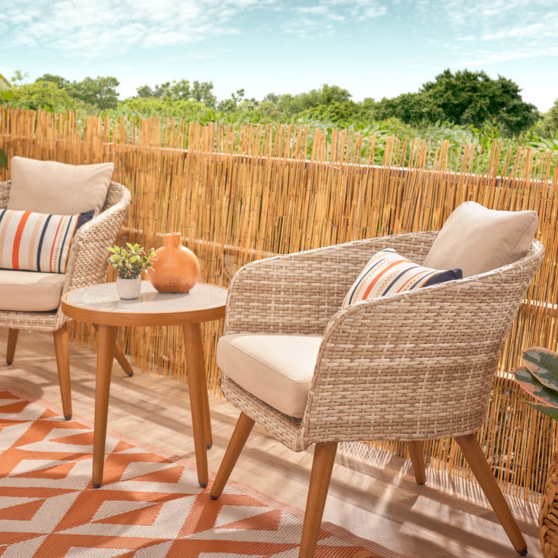 Juego de terraza de fibras naturales claras y cojines color crema, una mesa de madera redonda y una alfombra para exterior con patrones naranja
