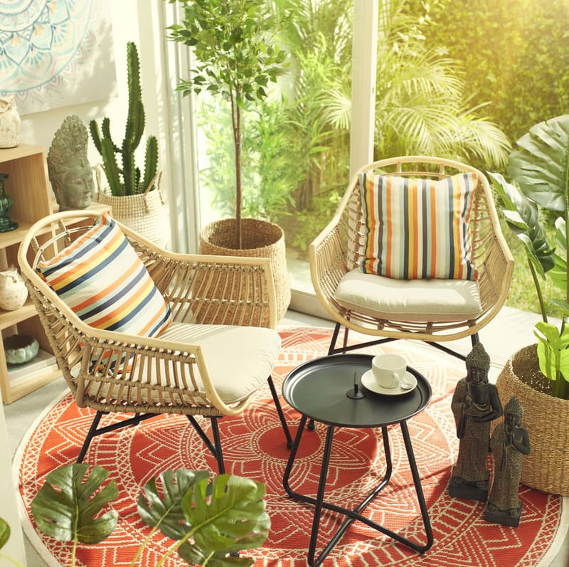 Espacio interior con dos sillas de terraza de fibras naturales con cojín color crema, una mesa de centro pequeña y redonda y macetas.