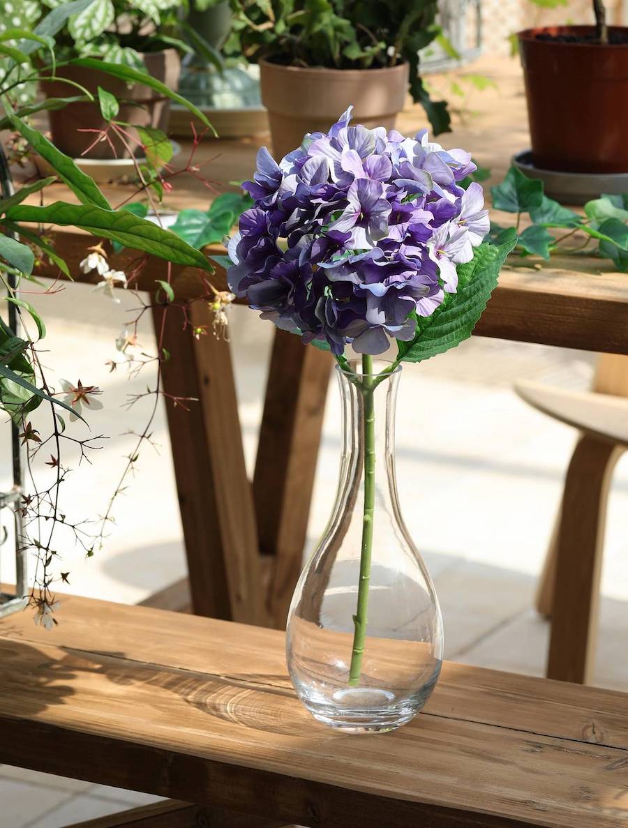 Detalle de una flor de hortensia lila artificial en un macetero de vidrio sobre una banca de madera.