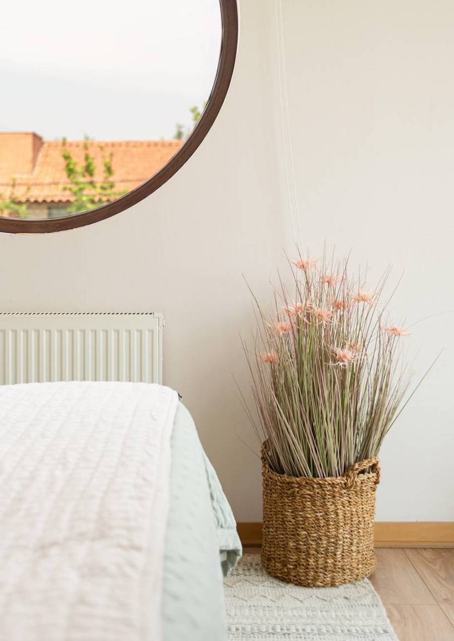 Rincón de un dormitorio con ropa de cama blanca y una planta artificial grass rosado verde dentro de un canasto de fibras naturales.