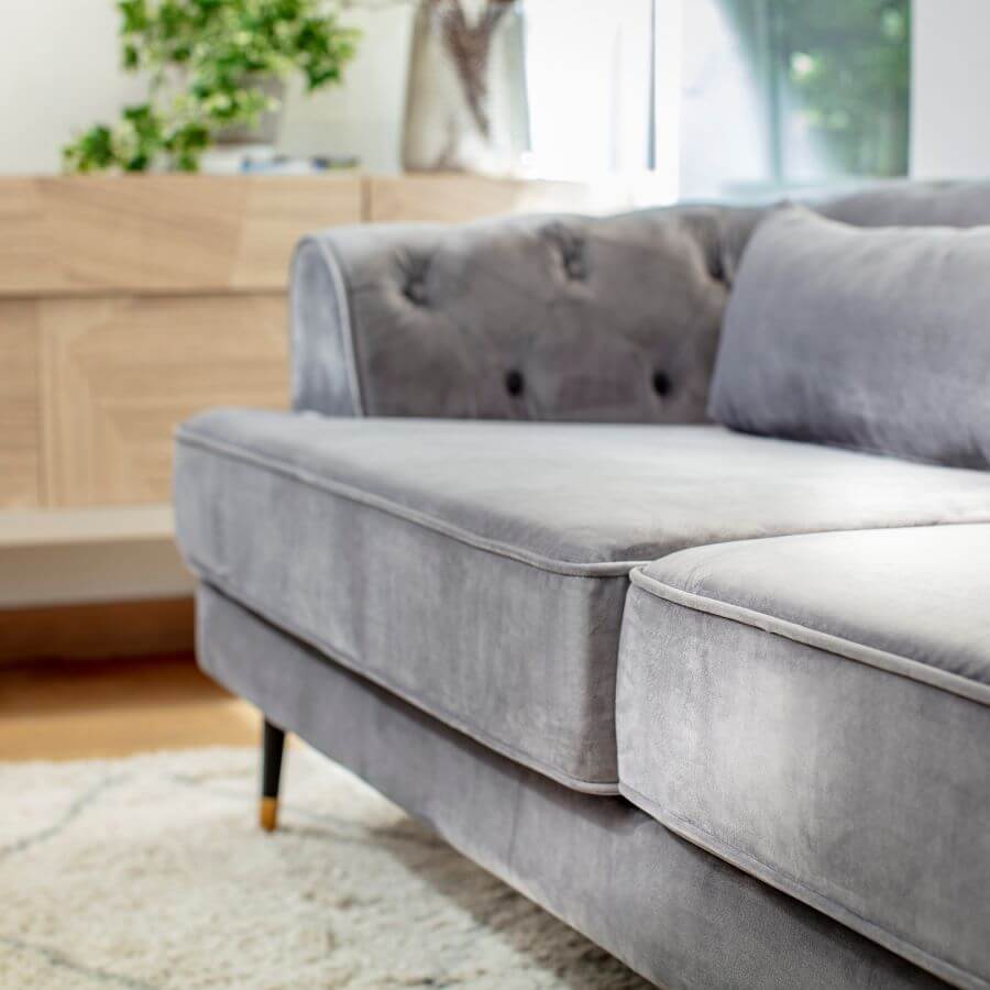 Detalle de sofá gris, de tela aterciopelada, con patas delgadas de color negro y dorado. Alfombra color beige con líneas grises