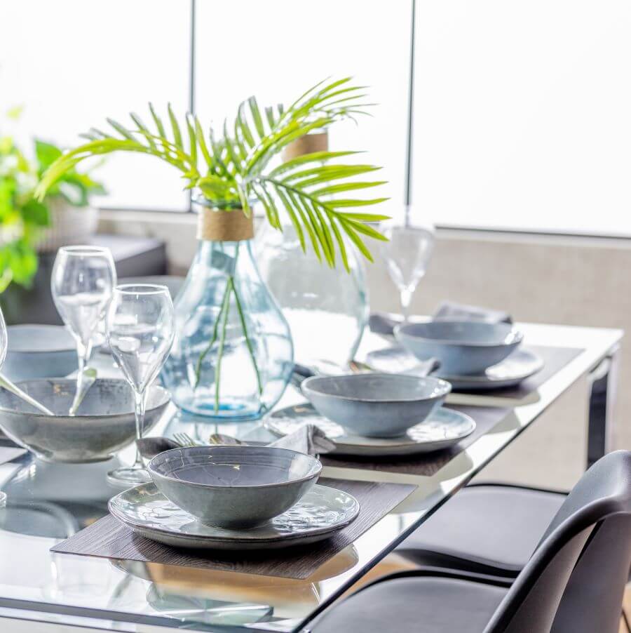 Mesa de comedor de vidrio, rectangular. Sobre ella hay puestos, con platos y bowls de cerámica color gris. Copas de vidrio y un gran florero de vidrio con hojas verdes como adorno.