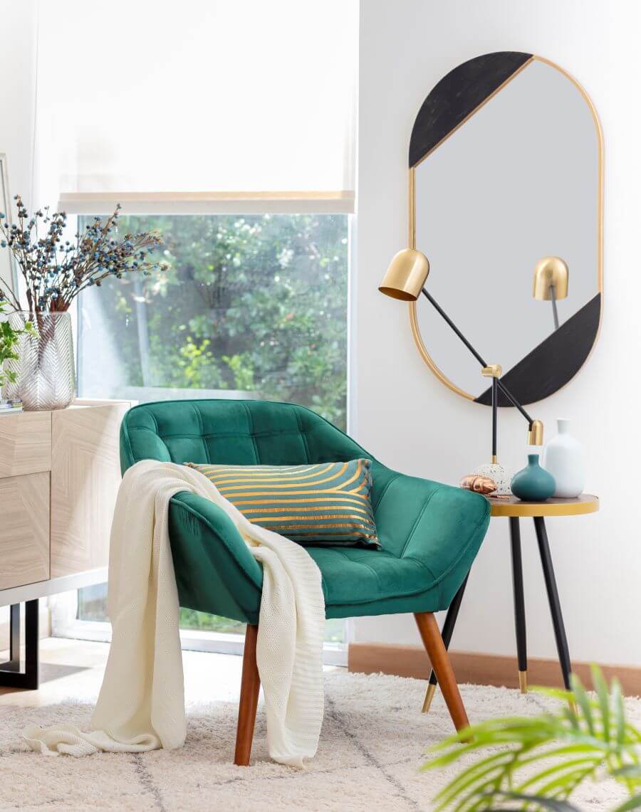 Sofá de color verde con patas de madrea, sobre él hay una manta beige y un cojín alargado. Mesa de apoyo pequeña, redonda, con patas de color negro. En el muro blanco hay un gran espejo ovalado con marco dorado.