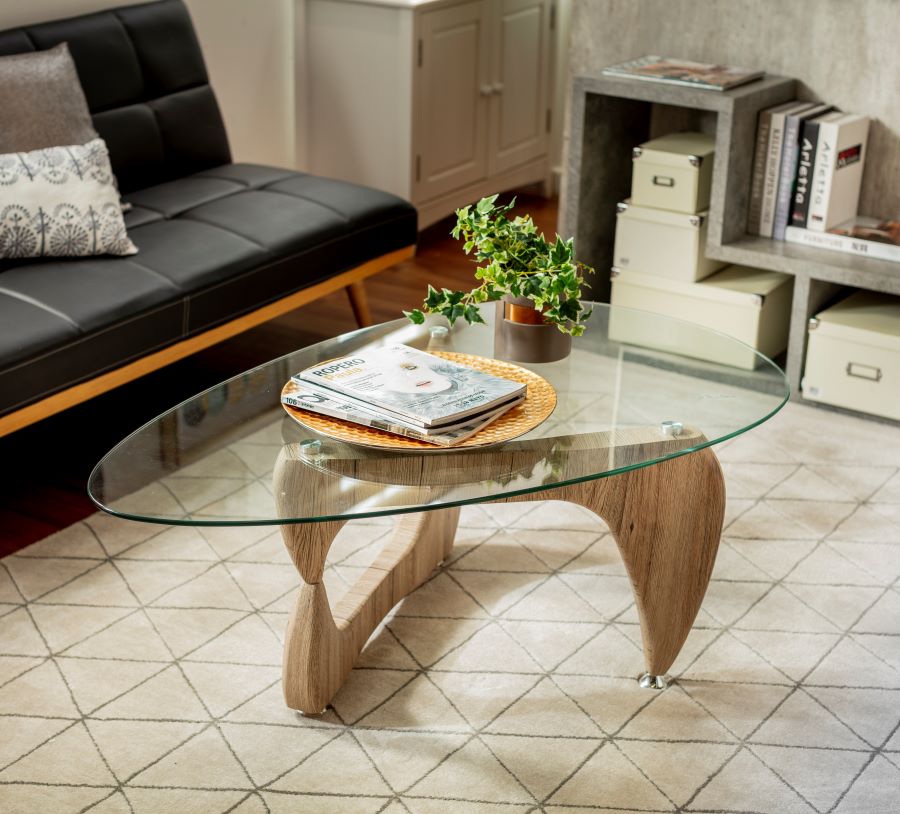 Mesa de centro con patas de madera y base de vidrio. Decorada con una bandeja redonda dorada con revistas y una pequeña planta en un macetero gris oscuro.