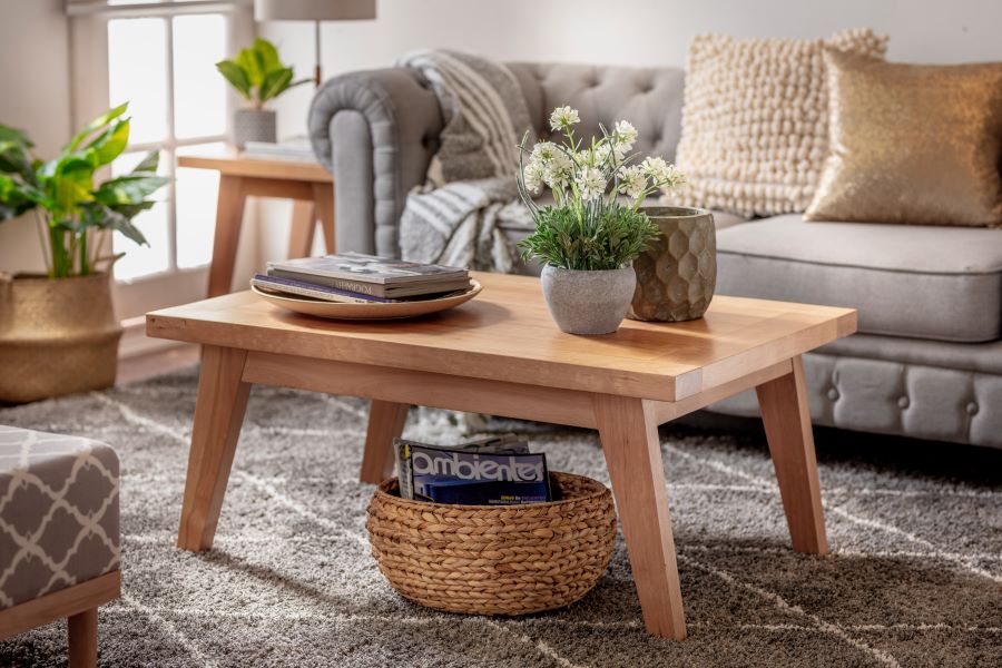 Mesa de centro de madera, de estilo clásico. Decorada con una bandeja con revistas, un macetero con flores blancas y un macetero gris. Bajo la mesa hay un canasto de mimbre con revistas.