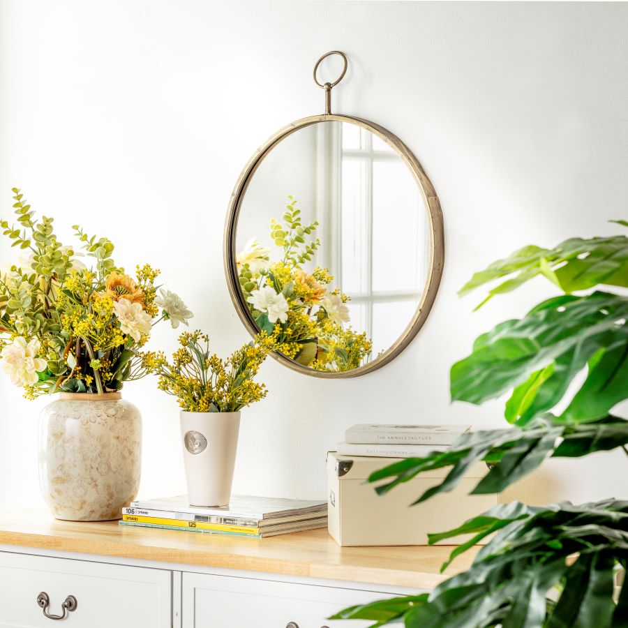 Recibidor pequeño con mueble blanco, espejo redondo y floreros con flores amarillas. Gran planta verde.