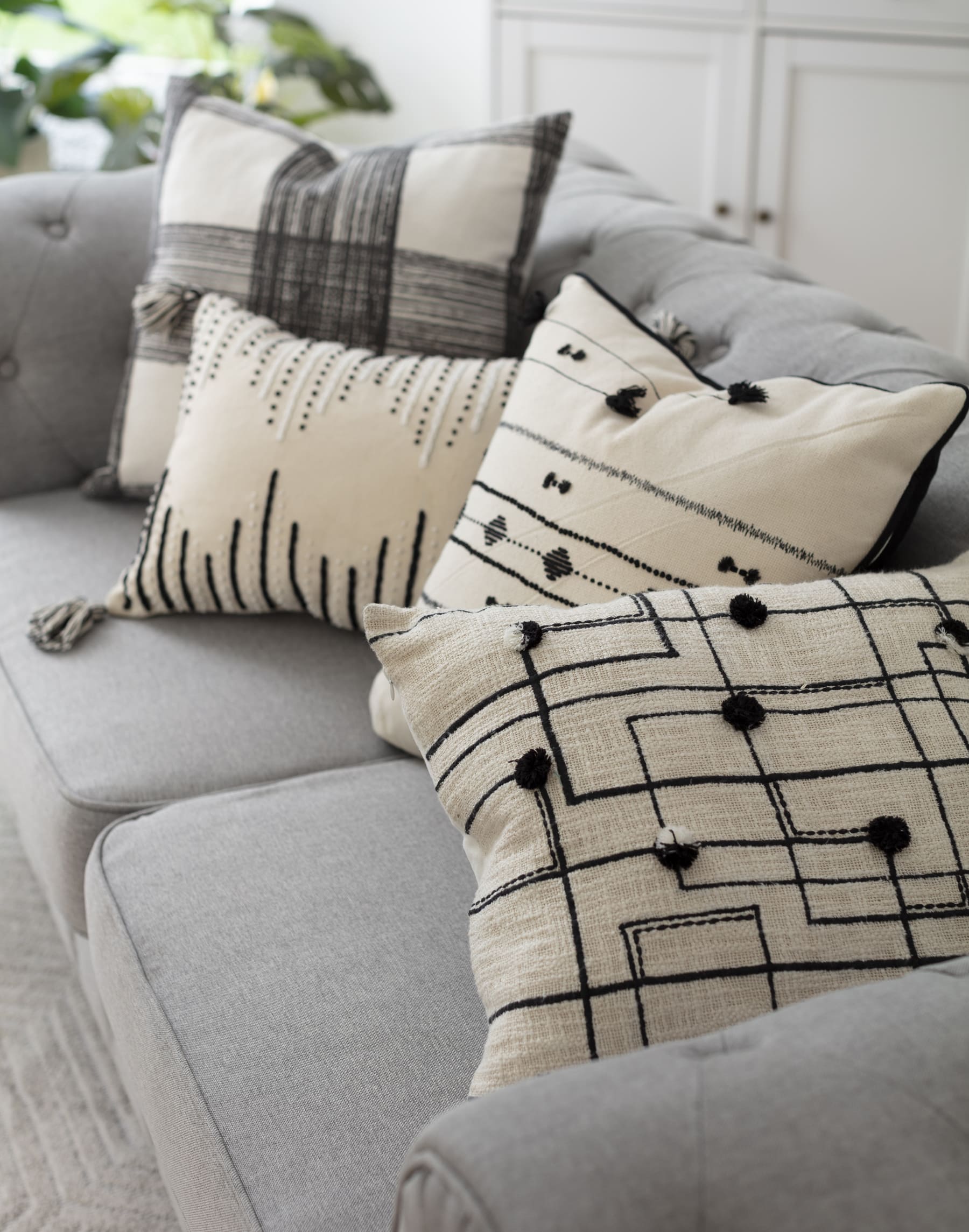 Cuatro cojines color crema con distintos patrones negros sobre un sofá gris con respaldo estilo capitoné.