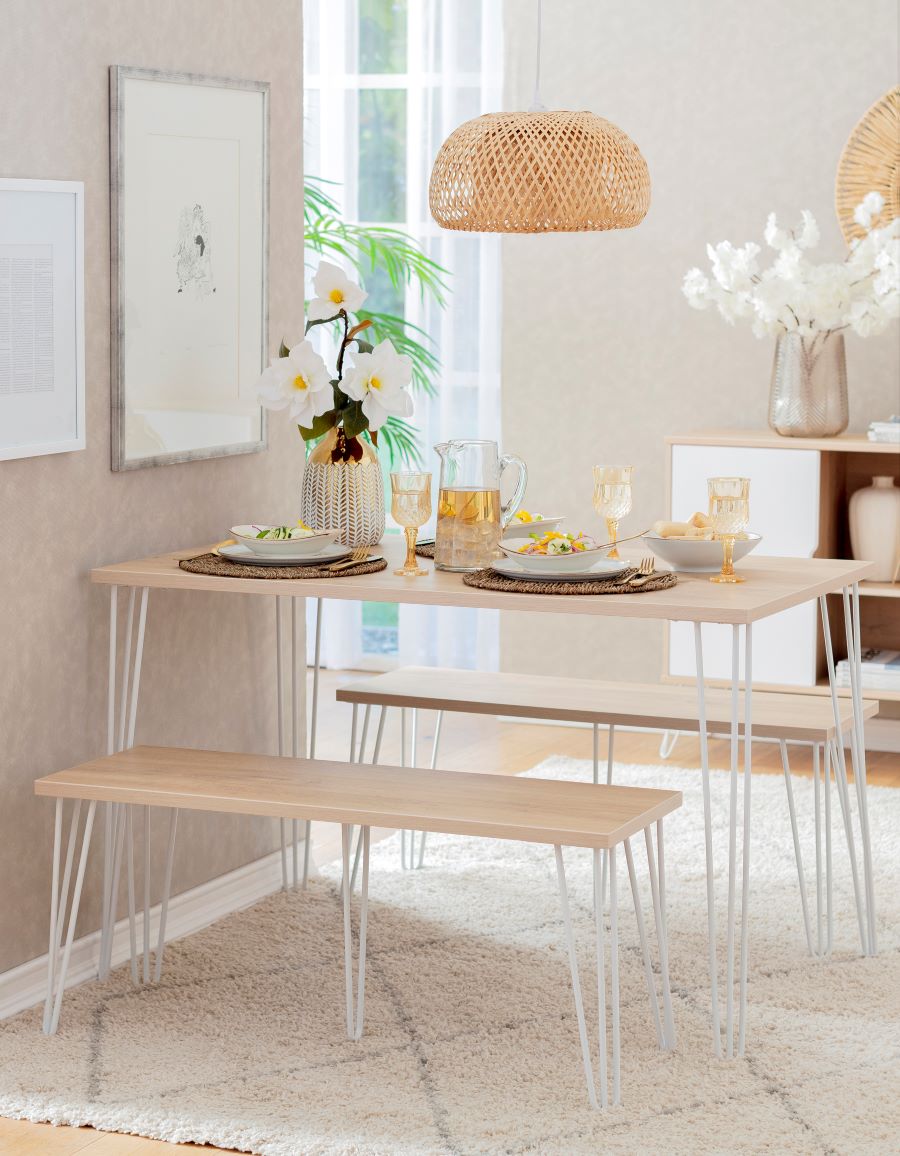 Living comedor pequeño y rectangular. Mesa rectangular de madera con patas de fierro blanco. Dos banquetas a juego. Lámpara de techo de mimbre, estilo boho.