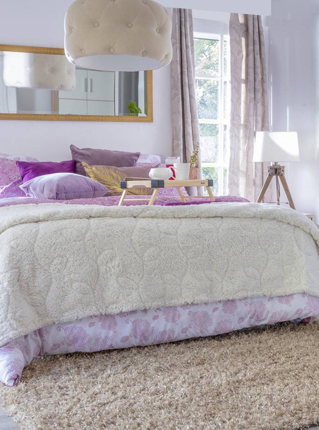 Dormitorio de paredes blancas con un gran espejo con marco dorado puesto horizontalmente sobre el cabecero de la cama. Las almohadas y cojines son morados, hay una gran manta blanca a los pies de la cama y en el suelo, una alfombra estilo shaggy beige.