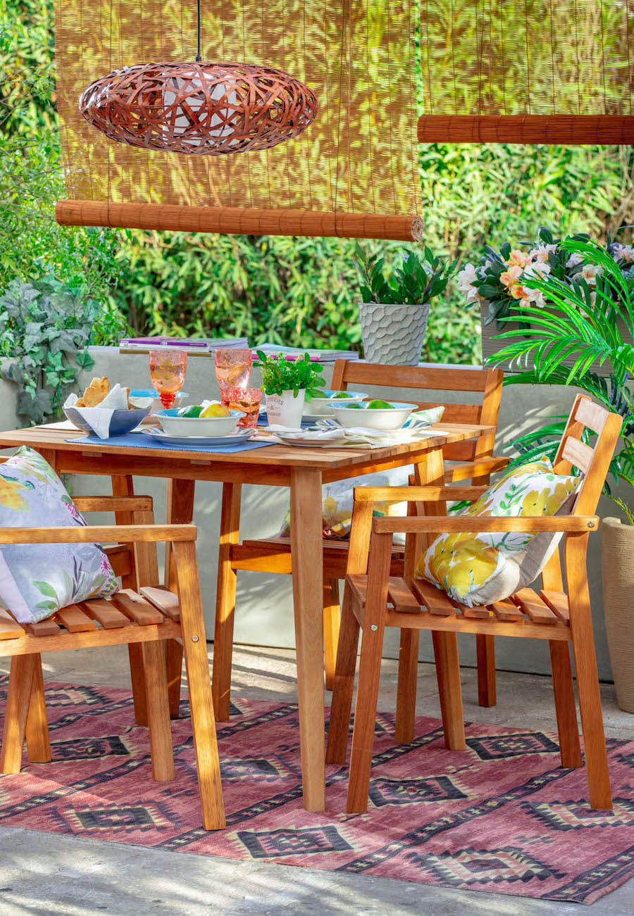 Terraza con cortinas enrollables de bambú para mayor privacidad. Mesa de comedor de madera con 4 sillas y vajilla de colores. Cojines con flores