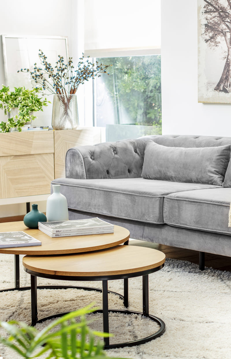 Living con un sofá gris velvet con respaldo estilo capitoné con una mesa de centro gemelar con cubierta redonda de madera y base de metal negro, uno es más grande que la otra.