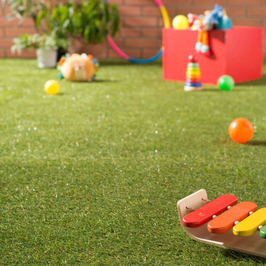 Detalle de terraza con pasto sintético. Sobre el pasto hay juegos infantiles, como pelotas y un xilófono. En la parte de atrás se ve un muro de ladrillo y plantas.