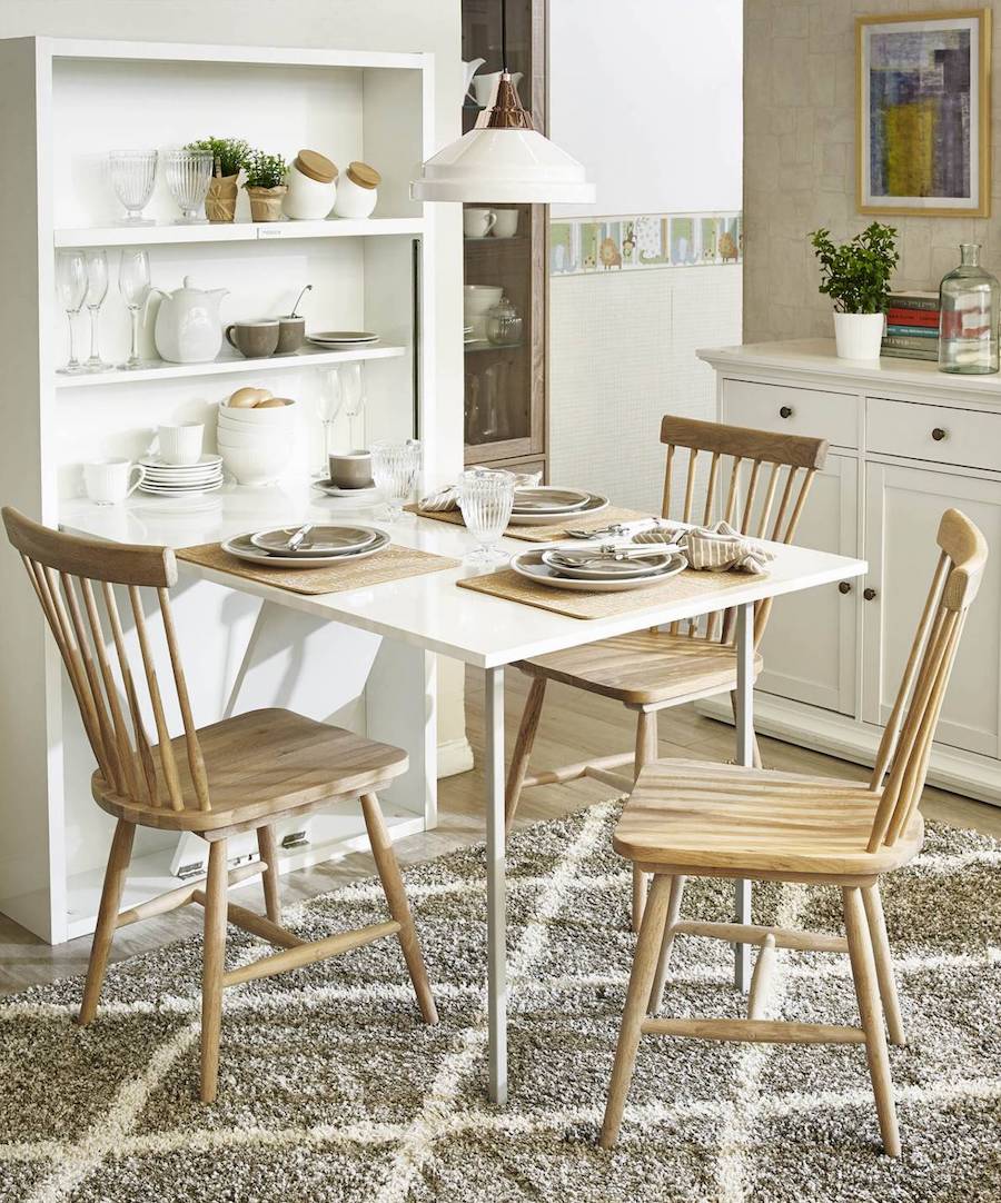 Cocina de estilo nórdico con un estante blanco que incluye una mesa abatible. Sobre ella hay platos, cubiertos, copas e individuales, y alrededor, 3 sillas de madera clara. En la parte del estante hay copas, plantas, frascos, tazas, platos y pocillos.