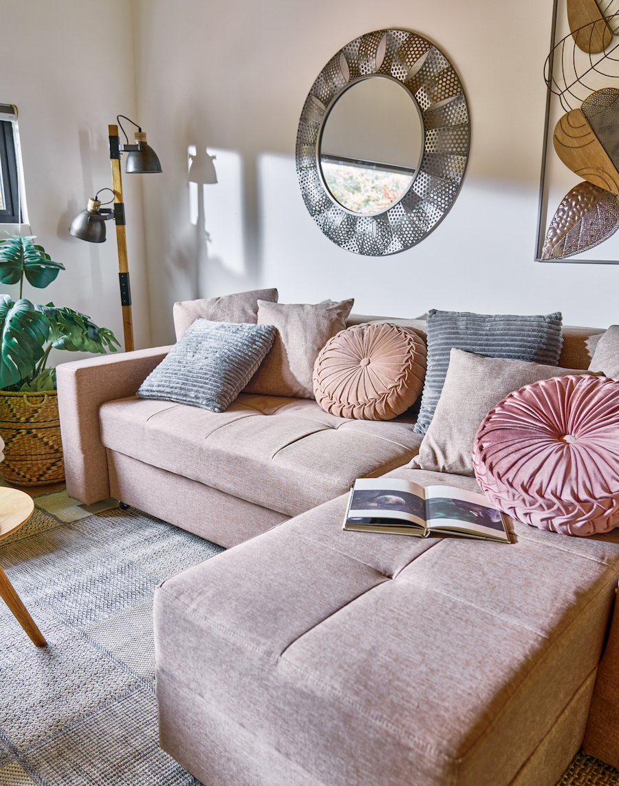 Sala de estar con un futón seccional beige con cojines en tonos beige, grises y rosados sobre él. En la pared del fondo hay un espejo redondo y un cuadro, ambos de estructura metálica.