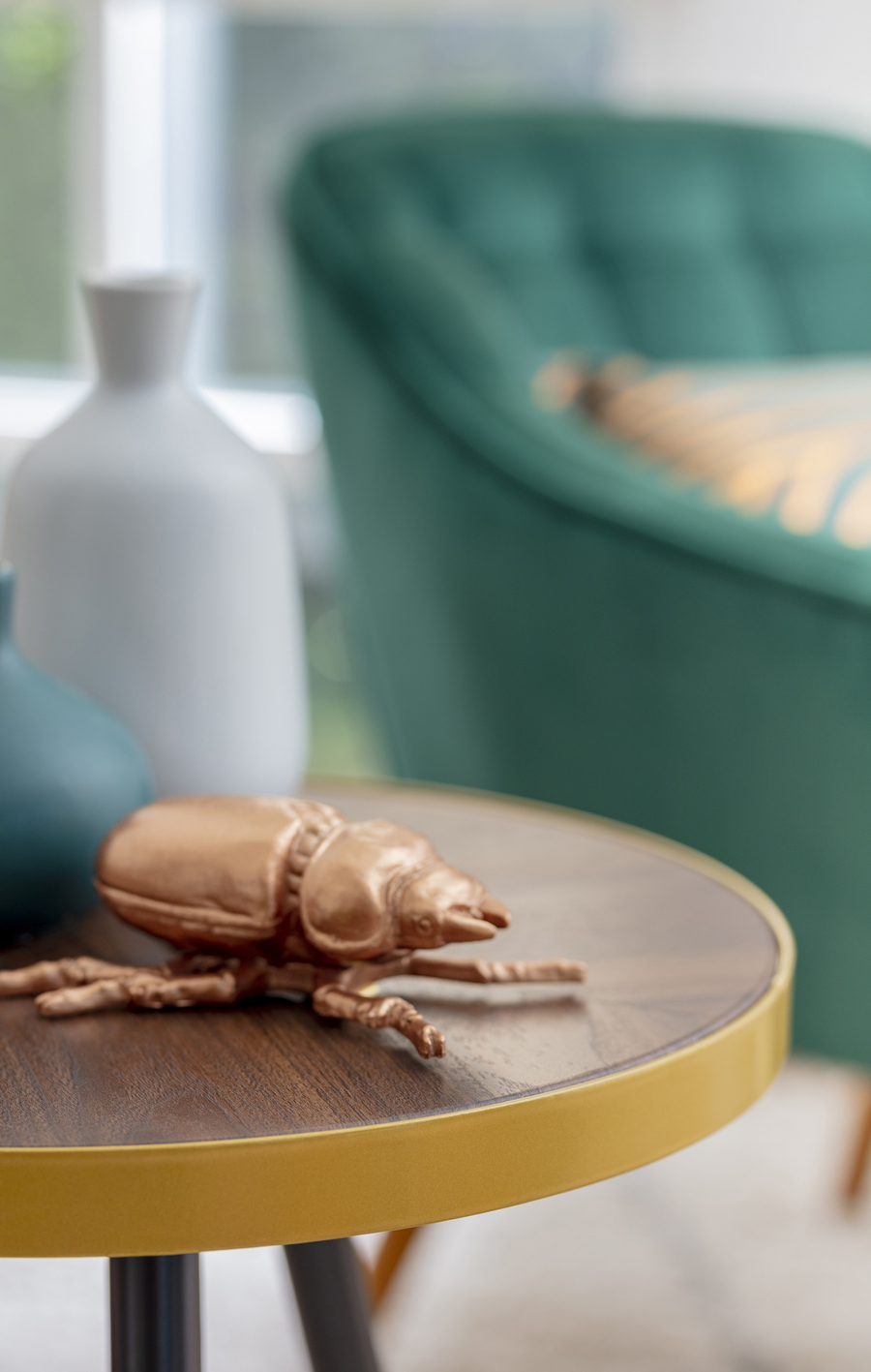Detalle de una mesa lateral de madera, sobre la cual hay un adorno de un escarabajo dorado. Atrás se ve un jarrón blanco y un sitial color verde