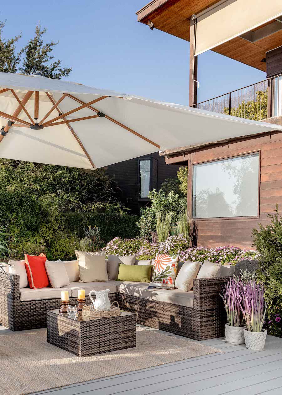 Terraza con gran quitasol y un juego de living exterior de ratán con cojines beige y otros de colores. Atrás se ve una gran casa de madera.