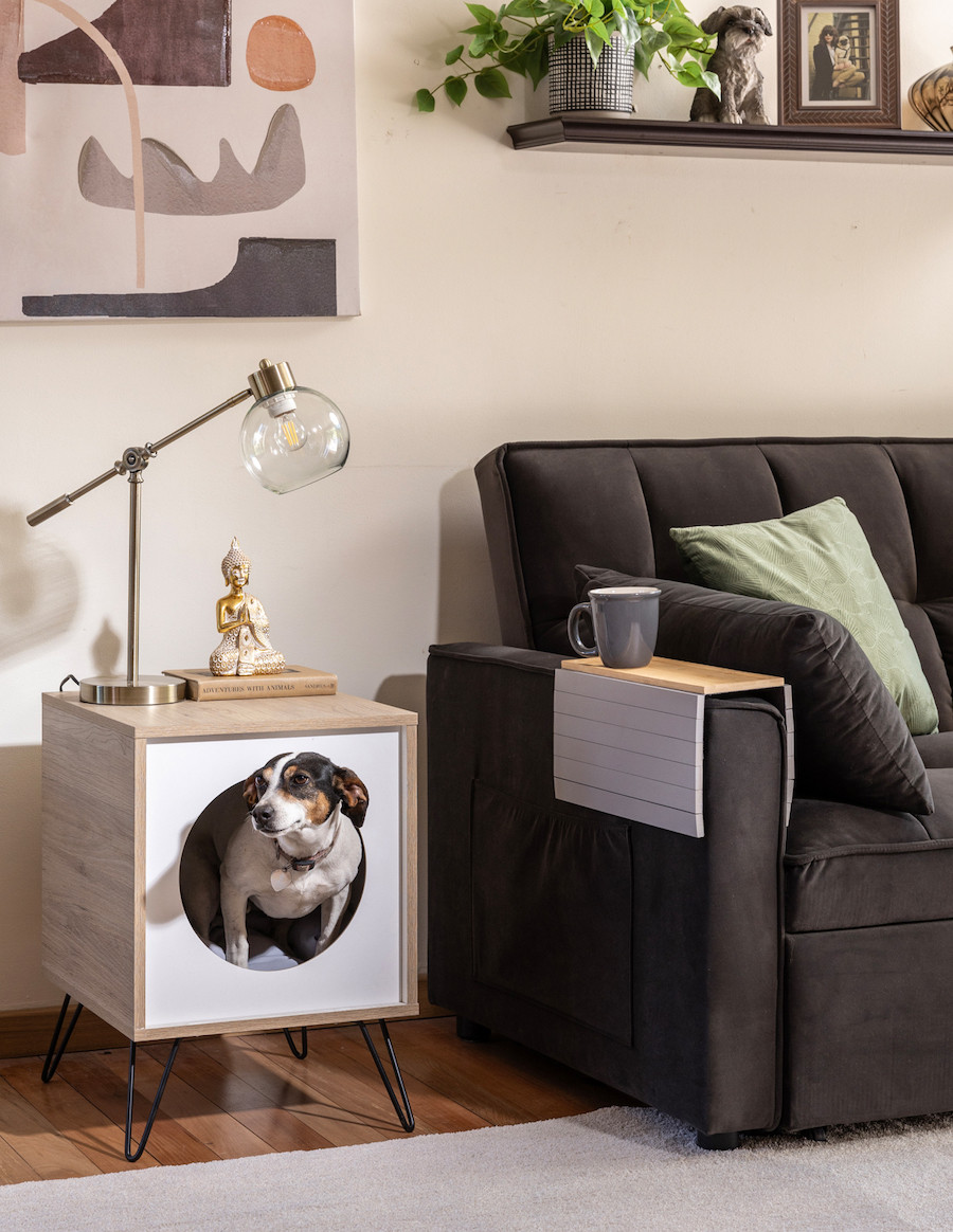 Mueble mutifuncional que mezcla una mesa latera con una casa para gatos, Es de madera, cuadrada con una puerta redonda. Está junto a un sillón negro.