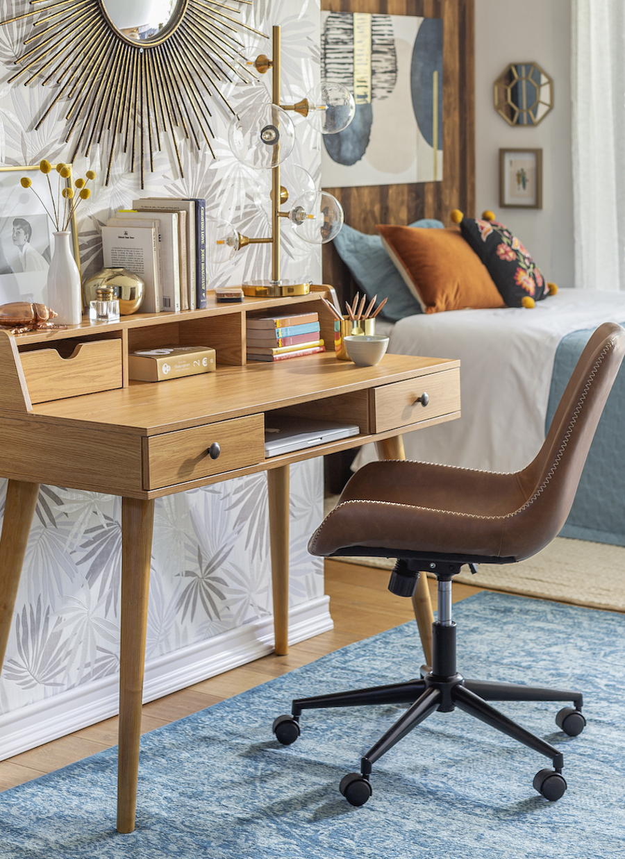 Escritorio dentro de un dormitorio. Escritorio de madera estilo vintage con silla café con ruedas. Alfombra azul y papel mural con figuras de hojas.