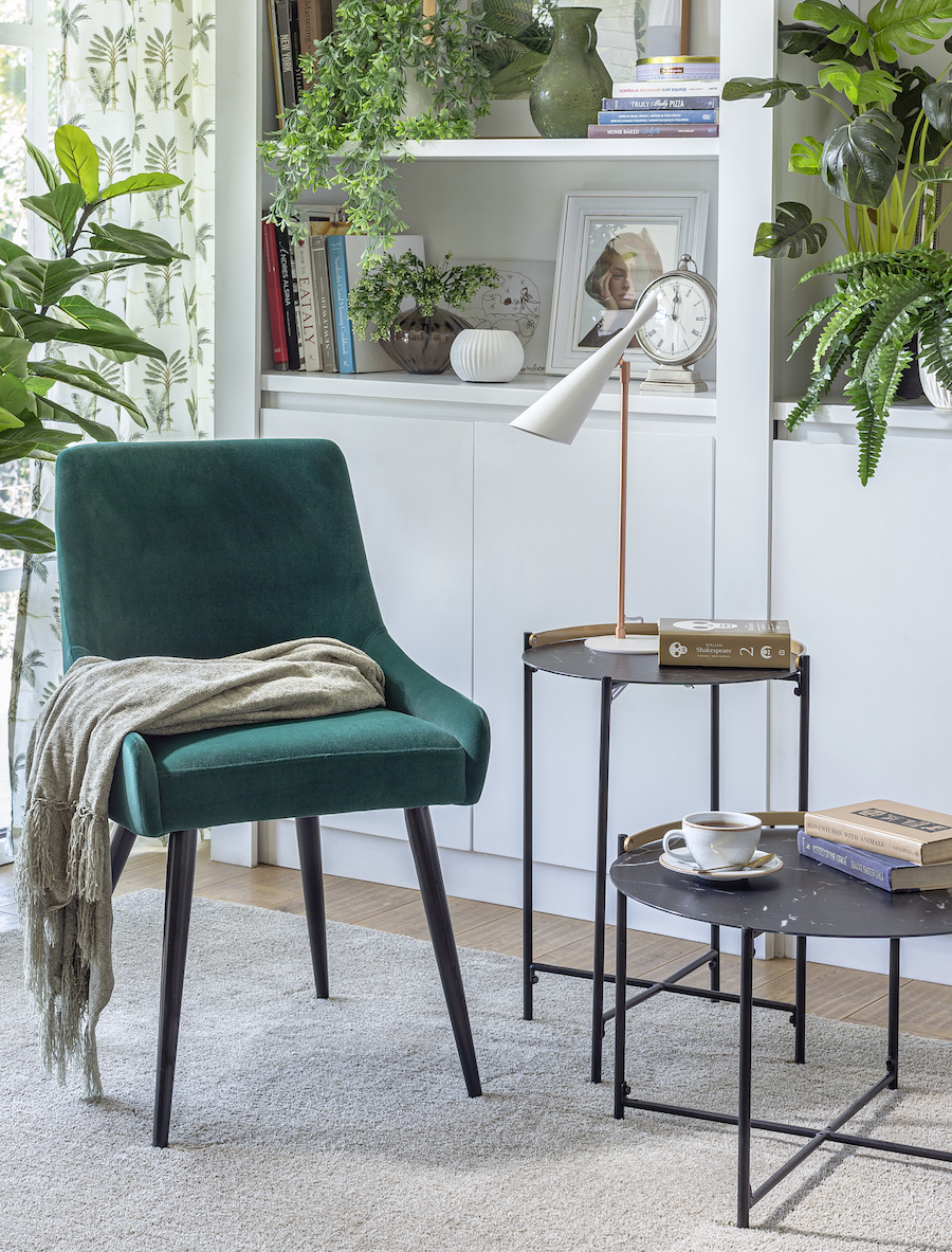 Rincón de un living con un sitial verde junto a un set de mesas negras. Alfombra beige y atrás un gran mueble con repisas color blanco.