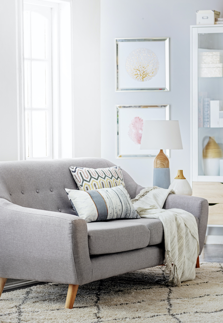 Living de estilo clásico con sofá gris con cojines y una manta. Junto a él hay una mesa de apoyo con una lámpara. Atrás hay una ventana y un muro con dos cuadros.