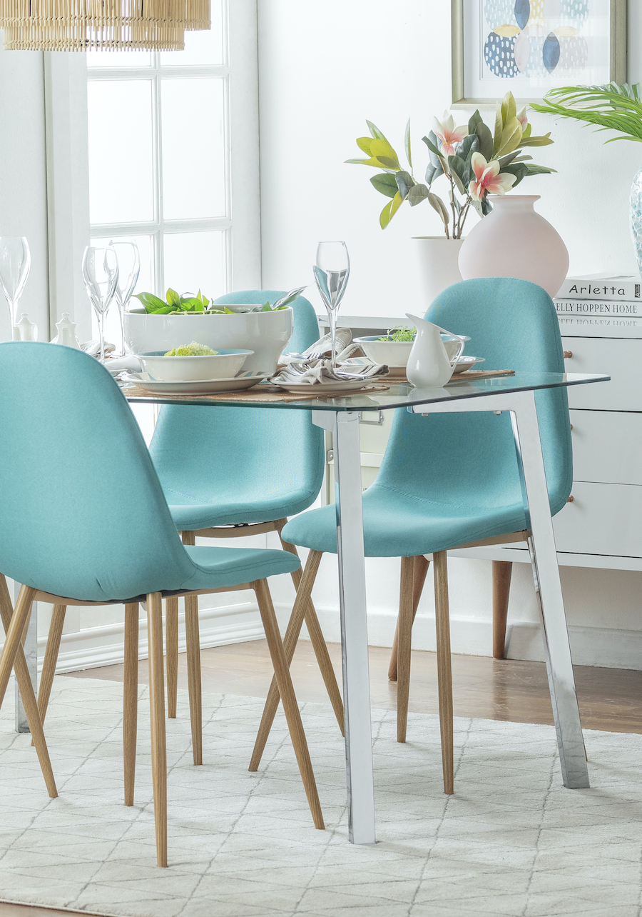 Comedor con mesa de vidrio y patas metálicas. Silla estilo eames color turquesa. Alfombra blanca y mueble de madera blanco en la parte posterior