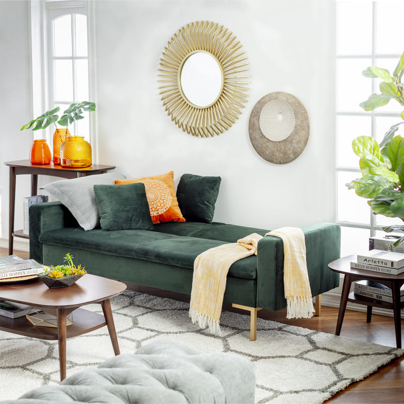 Living con un futón velvet verde oscuro, sobre una alfombra gris con patrones, que también sostiene una mesa de centro de madera oscura. También, en la pared, hay dos espejos redondos con marcos dorados.