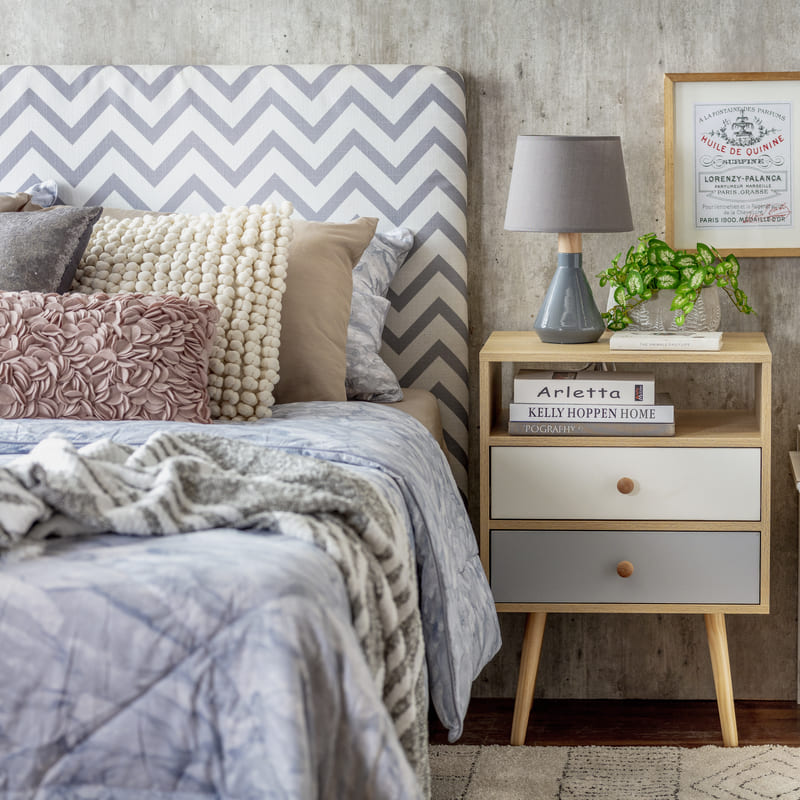 Dormitorio con un respaldo tapizado con una tela con patrón en zigzag, una ropa de cama azul y cojines de varios colores y estilo. Al lado, hay un velador de madera con dos cajones con distintos colores.