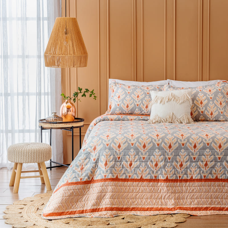 Dormitorio estilo Boho-chic, con una cama de dos plazas y un quilt con estampado celeste y rojo. Hay una mesa lateral, una banqueta, una lámpara y una alfombra de fibras naturales.