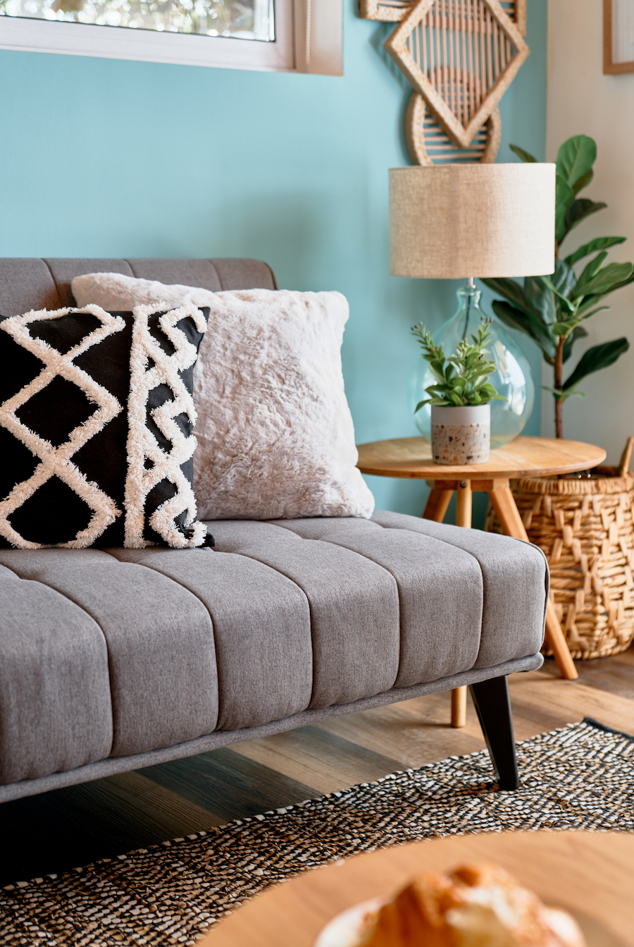 Futón gris con dos cojines, unos blanco peludo y otro negro con blanco estilo boho. Mesa de apoyo de madera, redonda, con lámpara de mesa de vidrio y pequeña planta. Pisos de madera y muro celeste